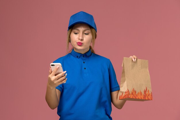 淡いピンクの食品パッケージを保持している彼女の電話を使用して青い制服を着た女性の宅配便、サービス制服の配達の仕事