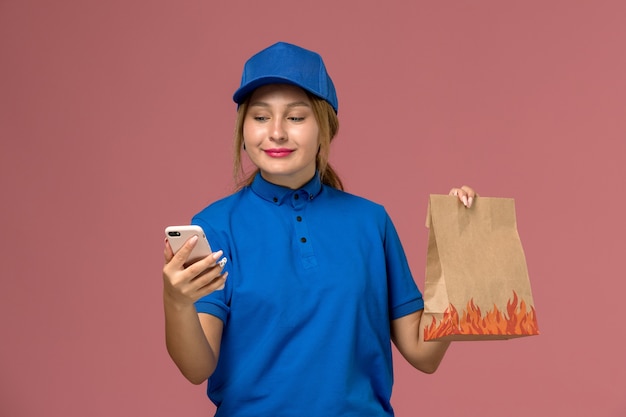 female courier in blue uniform using her phone holding food package with smile on light pink, service uniform delivery job