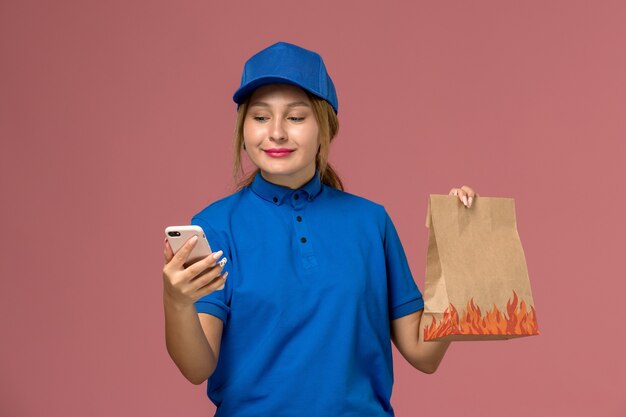 밝은 분홍색, 서비스 유니폼 배달 작업에 미소로 음식 패키지를 들고 그녀의 전화를 사용하여 파란색 유니폼 여성 택배