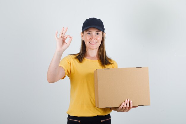 Женский курьер держит картонную коробку с футболкой, штанами, кепкой и выглядит радостно