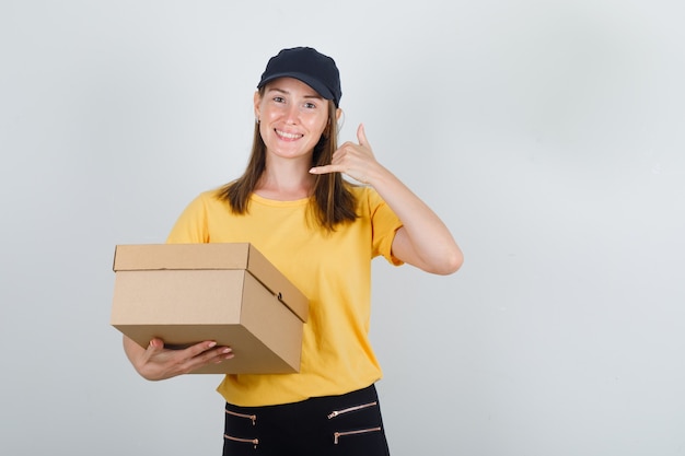 Female courier holding cardboard box with phone gesture in t-shirt, pants, cap and looking glad