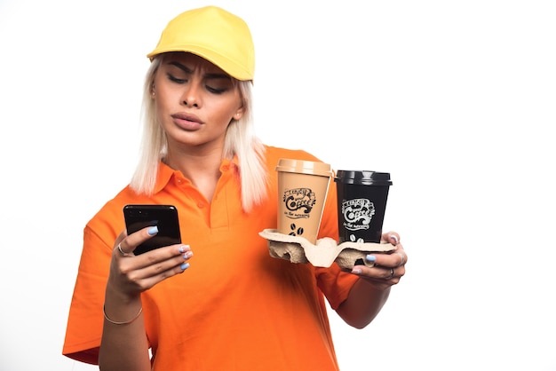 Женский курьер держит две чашки кофе на белом фоне при использовании телефона. Фото высокого качества