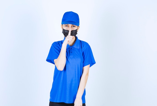 マスクと青い制服を着た女性の宅配便が口を閉じて沈黙を求めています。