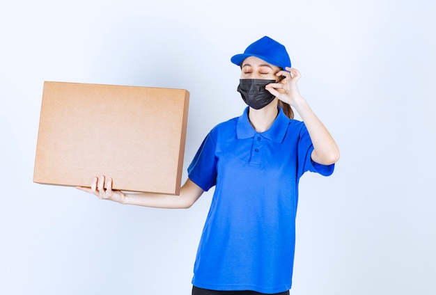 Foto gratuita corriere femminile in maschera e uniforme blu che tiene in mano un grosso pacco di cartone e mostra un segno positivo con la mano