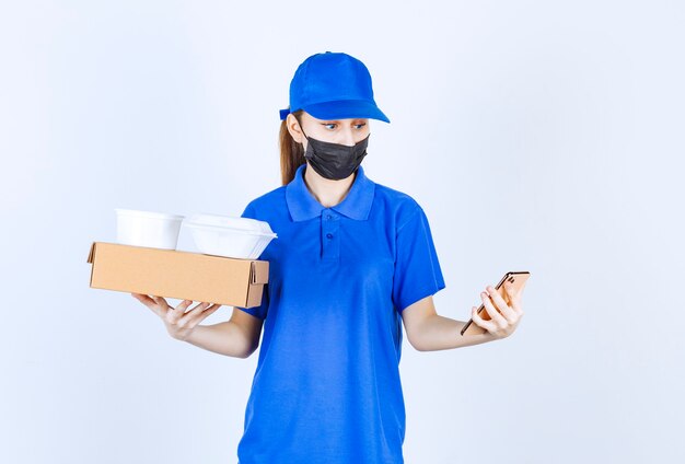 Женский курьер в маске и синей форме держит картонную коробку, пакеты на вынос и делает селфи