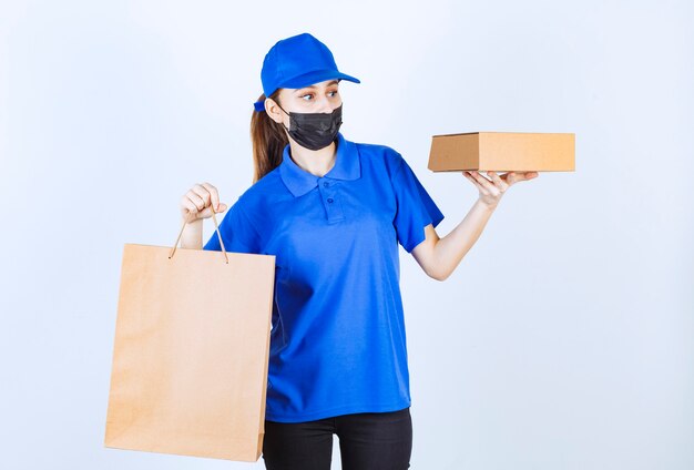 Female courier in mask and blue uniform holding a cardboard shopping bag and a box.