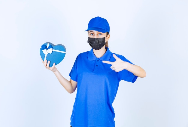 Female courier in mask and blue uniform holding a heart shape gift box