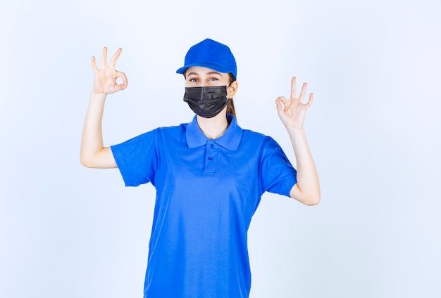 Free photo female courier in mask and blue uniform showing positive hand sign.