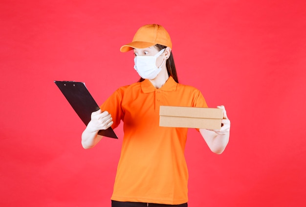 Free photo female courier in orange color dresscode and mask holding a cardboard box and reading the name and address