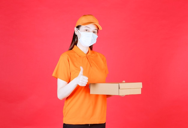 Female courier in orange color uniform and mask holding a cardboard box and showing positive hand sign.