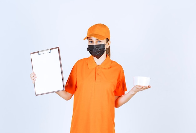 Corriere femminile in uniforme arancione e maschera nera con in mano un bicchiere di plastica e un elenco clienti