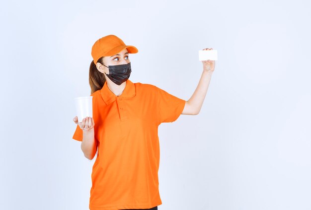 Female courier in orange uniform and black mask holding a plastic takeaway cup and presenting her business card