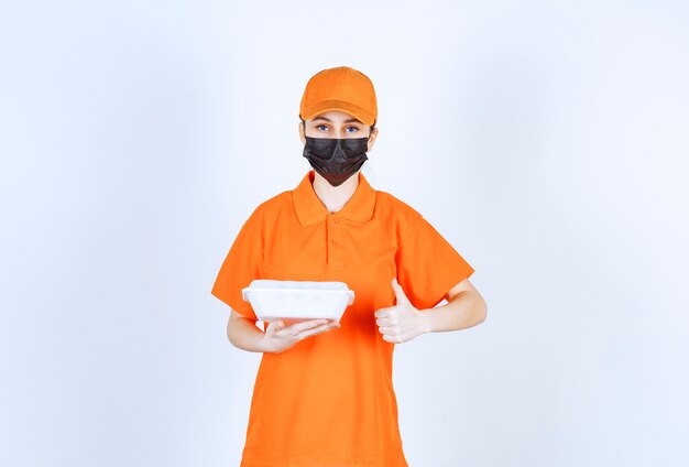Female courier in orange uniform and black mask holding a plastic takeaway food box and enjoying the taste