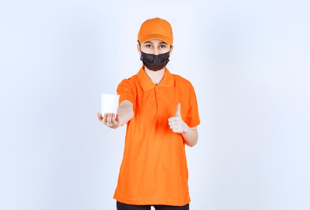 Female courier in orange uniform and black mask holding a takeaway drink and enjoying the taste