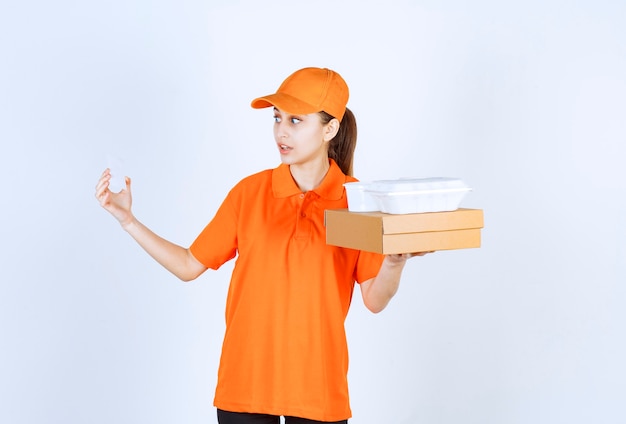 Female courier in orange uniform holding a cardboard box and a plastic takeaway box on it while presenting her business card.