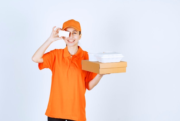 Female courier in orange uniform holding a cardboard box and a plastic takeaway box on it while presenting her business card.