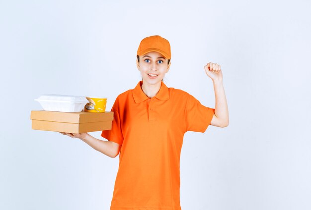 Female courier in orange uniform holding a cardboard box, a plastic takeaway box and a yellow noodles cup while showing positive hand sign.