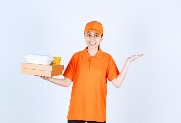 Corriere femminile in uniforme arancione con in mano una scatola di cartone, una scatola di plastica da asporto e una tazza di pasta gialla