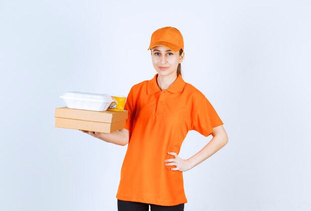 Курьер-женщина в оранжевой форме держит картонную коробку, пластиковую коробку для еды на вынос и желтую чашку с лапшой