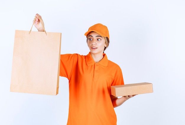 Female courier in orange uniform holding a cardboard parcel and a shopping bag