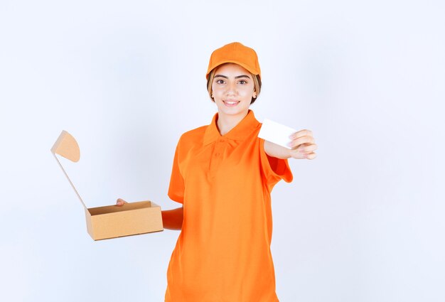 Female courier in orange uniform holding an open cardboard box and preseting her business card to the customer