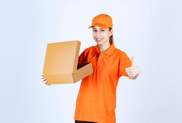 Female courier in orange uniform holding an open cardboard box and showing positive hand sign.