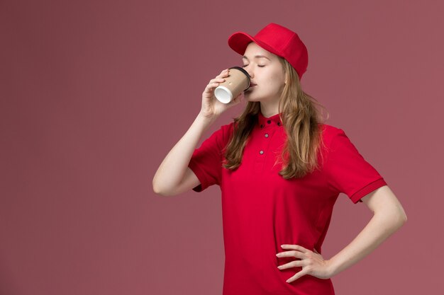 женщина-курьер в красной униформе пьет кофе на светло-розовом, рабочая форма службы доставки