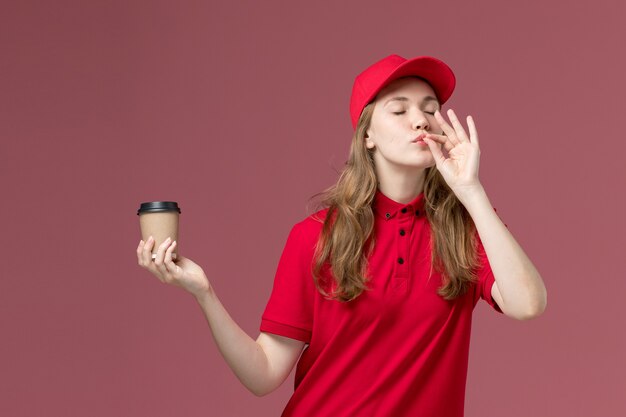 淡いピンクのおいしいサインを示す茶色のコーヒーカップを保持している赤い制服を着た女性の宅配便、仕事の制服サービスワーカーの配達