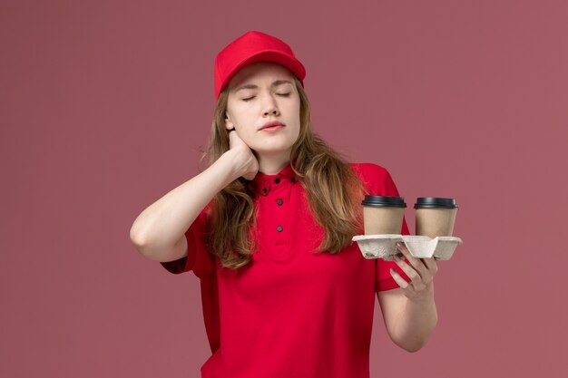 female courier in red uniform holding brown coffee cups having neck ache on pink, uniform service delivery worker job