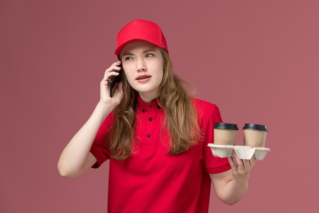 female courier in red uniform holding brown coffee cups and talking on phone on pink, uniform service delivery worker job