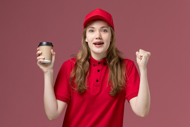 женщина-курьер в красной форме держит коричневую кофейную чашку доставки на светло-розовом, рабочая форма службы доставки