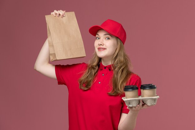 female courier in red uniform holding brown delivery coffee cups and food package on pink, uniform service delivery job worker