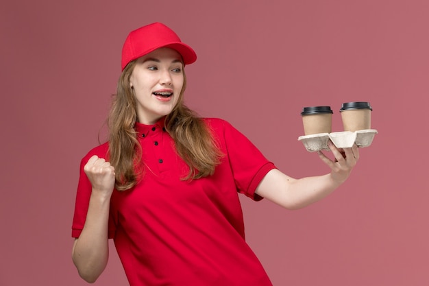 женщина-курьер в красной форме держит коричневые кофейные чашки на розовом, униформе служащий службы доставки