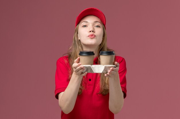 분홍색, 균일 한 직업 서비스 노동자에 공기 키스를 보내는 갈색 배달 커피 컵을 들고 빨간 제복을 입은 여성 택배
