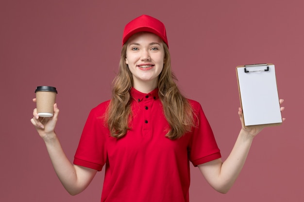 женщина-курьер в красной форме держит коричневый кофе доставки и блокнот с улыбкой на розовом, рабочая форма службы работника