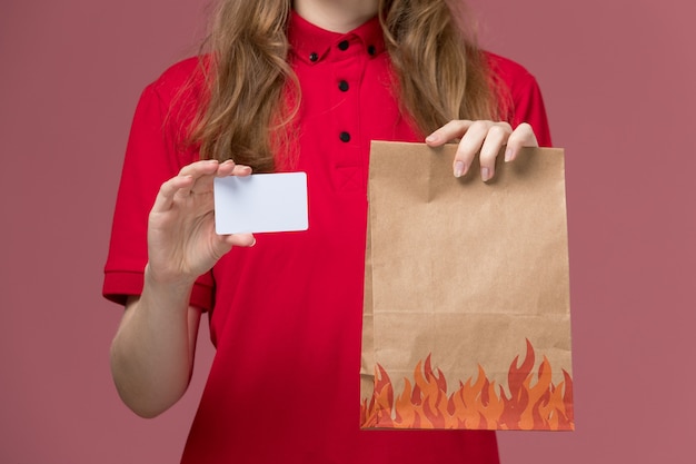 женщина-курьер в красной форме держит карточку и пакет с едой на розовом, униформе работник службы доставки