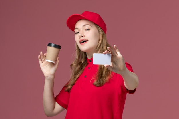 female courier in red uniform holding coffee cup along with white card on light pink, job uniform worker service delivery