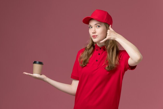 淡いピンクの、仕事の制服のサービスワーカーの配達でポーズをとってコーヒーカップの電話を保持している赤い制服を着た女性の宅配便