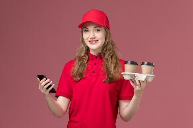 female courier in red uniform holding coffee cups and her phone on pink, uniform service delivery job