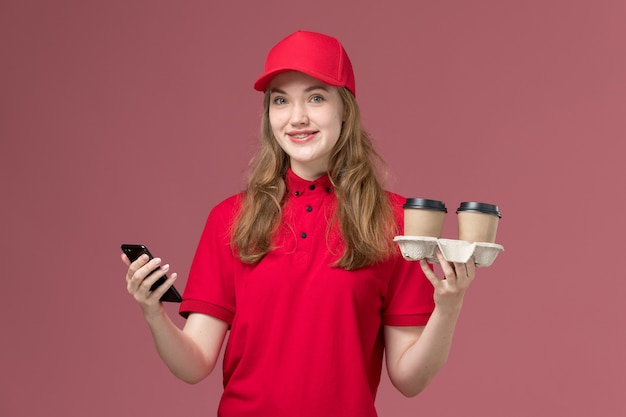 female courier in red uniform holding coffee cups and her phone on pink, uniform service delivery job