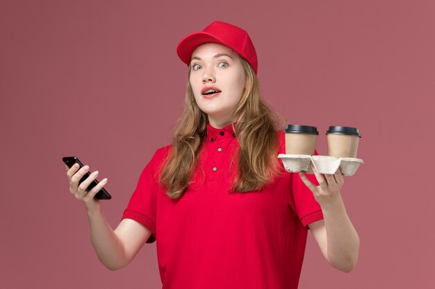ピンクの制服のサービス提供の仕事でコーヒーカップとスマートフォンを保持している赤い制服の女性の宅配便