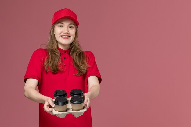 female courier in red uniform holding coffee cups and smiling on pink, uniform job worker service delivery