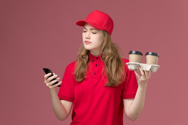 женщина-курьер в красной форме с доставкой кофейных чашек и телефоном на светло-розовом, униформе работница службы доставки