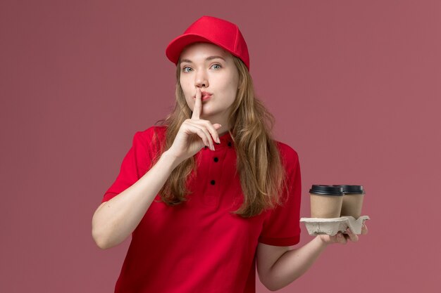 female courier in red uniform holding delivery coffee cups showing silence sign on light pink, job uniform service worker delivery