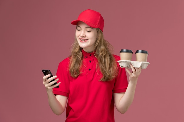 female courier in red uniform holding delivery coffee cups with phone on light pink, uniform service worker delivery