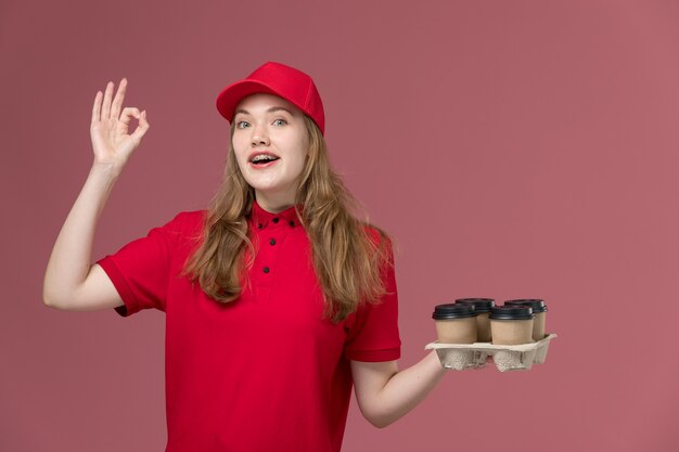 женщина-курьер в красной униформе, держащая кофейные чашки с улыбкой и знаком хорошо на розовом, рабочая форма работника службы доставки