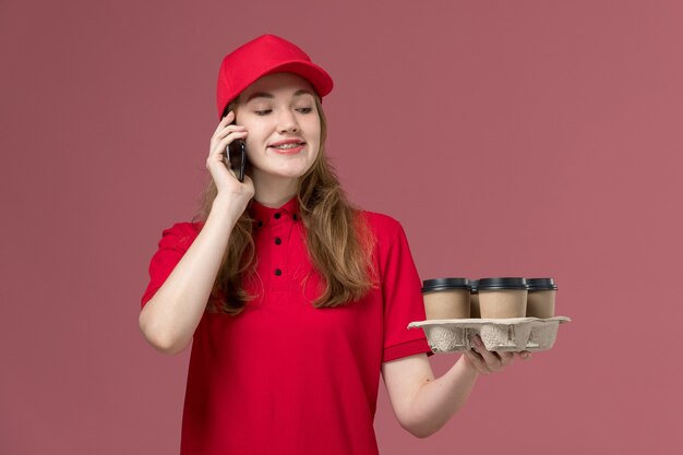 женщина-курьер в красной форме, держащая доставку кофе, разговаривает по телефону на розовом, служба доставки рабочей формы