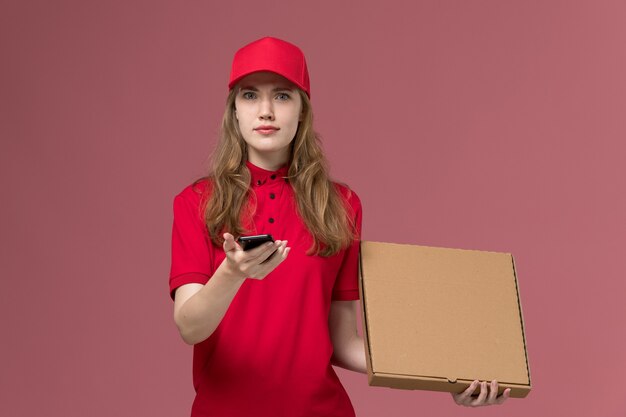 female courier in red uniform holding delivery food box and phone on light pink, uniform job service worker delivery girl