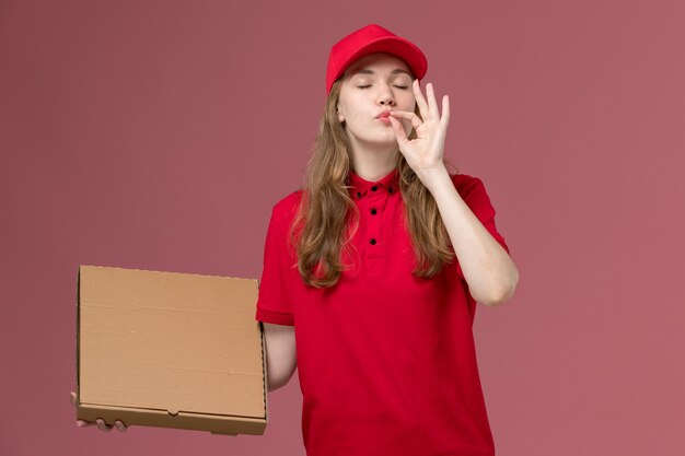 женщина-курьер в красной форме держит коробку с едой на розовом, униформенный работник службы доставки