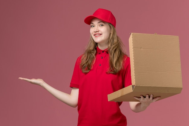 женщина-курьер в красной форме держит коробку с едой и улыбается на розовой, униформе службы доставки